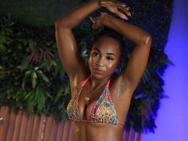 NathyRoss with shiny brown skin, small colorful bikini top showing her tits, arms raised, looking down, blue light in background, green plant on left, wooden fence below.