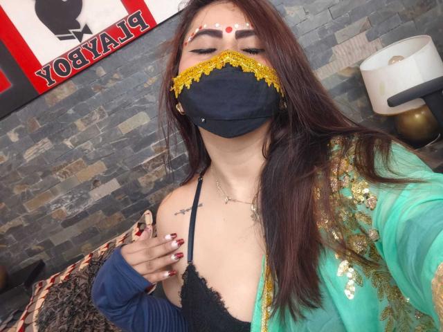 Dalia Dubey, con lunghi capelli castani, indossa una maschera nera con pizzo giallo, una maglia verde con paillettes dorate, un reggiseno nero in pizzo, tiene una mano sul seno e ha lo smalto rosso sulle unghie..