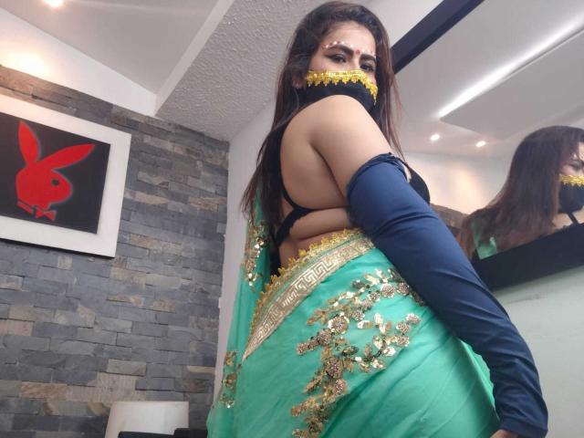 DaliaDubey with green saree, gold embroidery, black bra, navy elbow gloves, yellow mask, long dark hair, looking back, showing her ass, in a modern room.