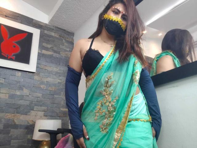 Dalia Dubey, con i capelli castani, indossa un reggiseno di pizzo nero, un sari verde con ricami dorati, bracciali blu, una maschera nera, ed è in piedi in una stanza. Uno specchio riflette la sua figura..