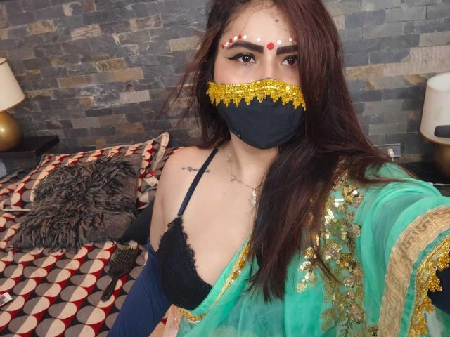 DaliaDubey with long brown hair, wearing a black lace bra, green and gold sequined shawl, black mask with gold trim, red dots on forehead, and tattoos on chest.