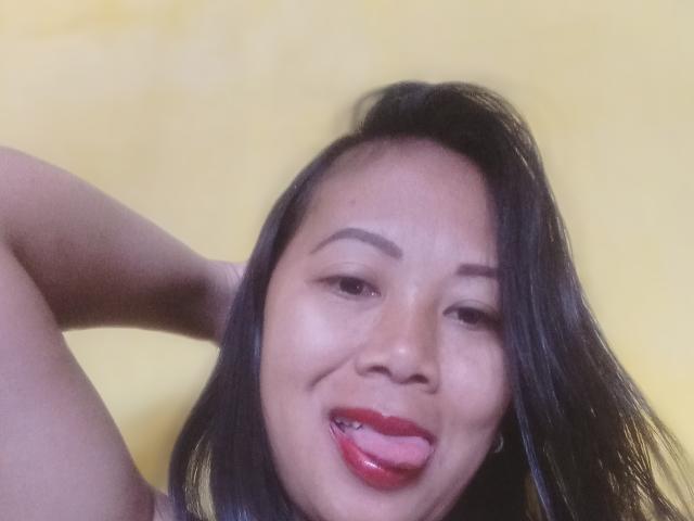 SarahFontaineX with black hair, tongue out, red lipstick, and raised arm, showing her slightly chubby face and small, cute nose, against a yellow background.
