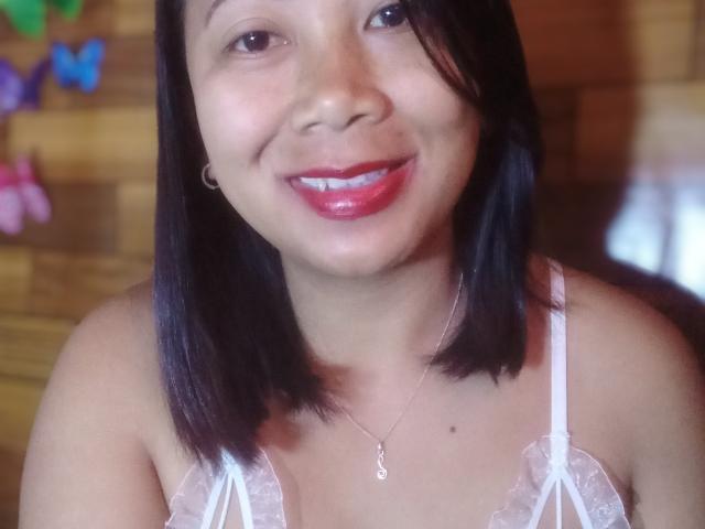 SarahFontaineX with dark brown shoulder-length hair, smiling with red lipstick, wearing a white lace bra, small silver necklace, and earrings, showing her tits.