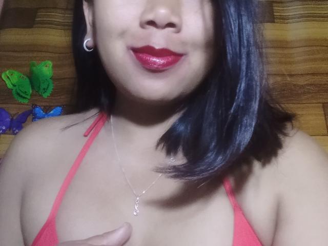 SarahFontaineX, com cabelo preto brilhante, batom vermelho e colar de prata, vestindo um top de biquíni vermelho, tem um rosto bonito e seios pequenos, em frente a uma parede de madeira..