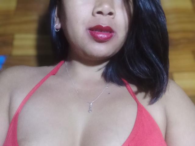 SarahFontaineX with red halter top, showing her large tits, red lipstick, black shoulder-length hair, silver necklace, and a small hoop earring, in a slightly blurred selfie.