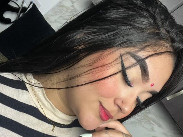 AnaShalimarr tem cabelos pretos e brilhantes, delineador preto marcante, pálpebras verdes, batom rosa, um bindi vermelho e está sorrindo com a mão perto da boca. Ela veste uma blusa listrada..