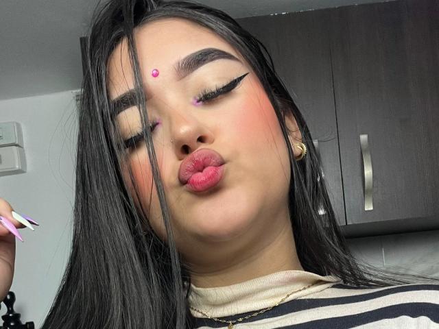 AnaShalimarr with long black hair, pink forehead dot, winged eyeliner, pink lipstick, pouty lips, holding pink and white nail polish, wearing striped shirt, in a kitchen.