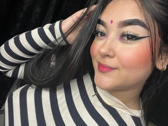 AnaShalimarr hat schwarze Eyeliner-Akzente, rosigen Rouge, roten Lippenstift und einen roten Punkt auf ihrer Stirn. Sie trägt ein gestreiftes Hemd und hat eine Hand in ihrem Haar..