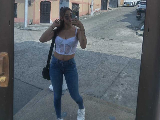 SashaHans, avec de grandes seins, porte un soutien-gorge en dentelle blanche, un jean bleu et des baskets blanches, et prend un selfie devant un miroir, debout sur un trottoir, tenant un téléphone, avec un sac noir..