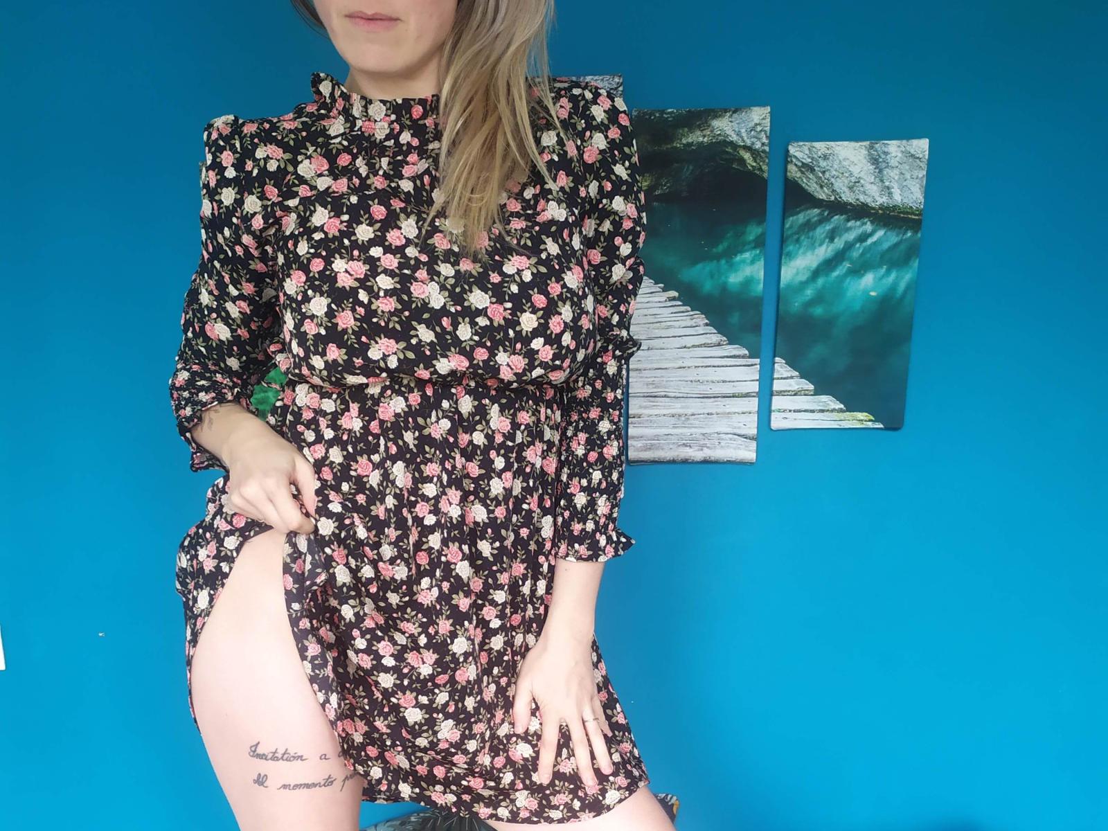 CassieReload portant une robe fleurie, tirant sur le côté pour montrer un tatouage sur la cuisse, avec des cheveux blonds, devant un mur bleu, avec deux tableaux représentant de l'eau turquoise en arrière-plan..