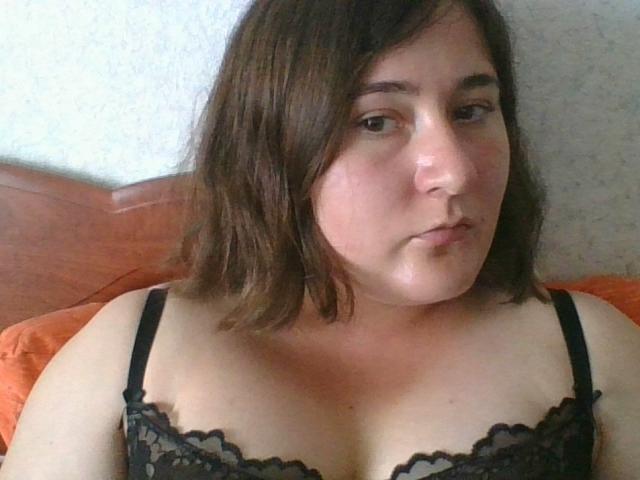 LadyDaniela with medium brown hair, wearing a black lace bra, looking slightly bored. Her tits are visible, showing a bit of cleavage. She's sitting on an orange pillow.