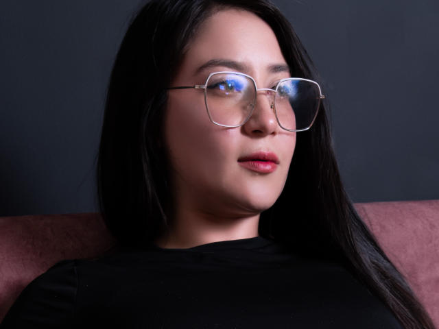 LeahMuniz with long black hair, wearing clear glasses and a black shirt, has pink lips and a subtle smile, sitting on a pinkish couch.