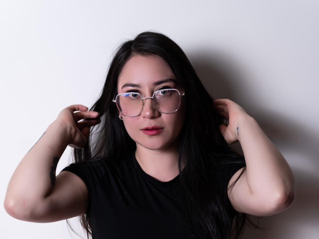LeahMuniz with long black hair, wearing clear glasses, and a black t-shirt, adjusting her hair with both hands, has pale skin and pink lips, looking straight at the camera.