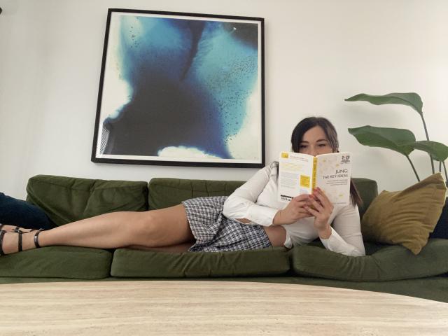 Dionisia, laying on green couch, wearing white long-sleeve shirt and checkered skirt, reading book, legs stretched out, brown hair, black heels, green pillow, yellow pillow, blue wall art.