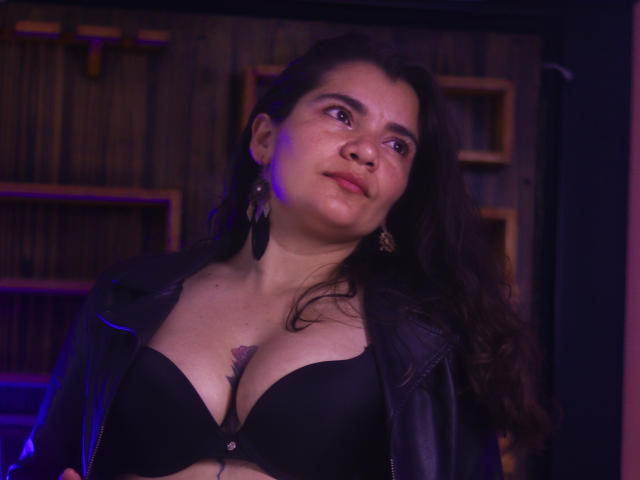 BulmaHot with medium tits in a black bra, wearing a leather jacket, has long dark hair, and a tattoo below her tits, looking up with a slight smile.