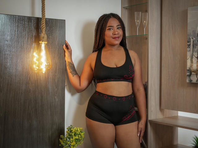 EbonyAmazing stands in black sports bra and shorts with red accents, showing her curvy ass and small tits, braided hair, tattoo on left arm, smiling at a light bulb.