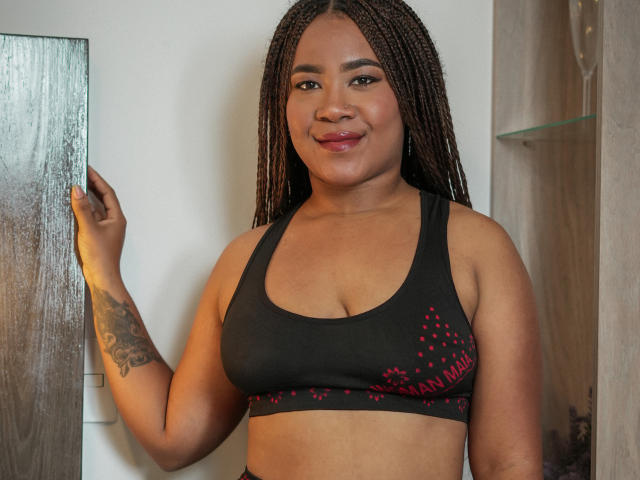 EbonyAmazing with braided hair, dark skin, and a tattoo on her right arm, wearing a black sports bra with red speckles, smiling slightly. She's holding a door frame.