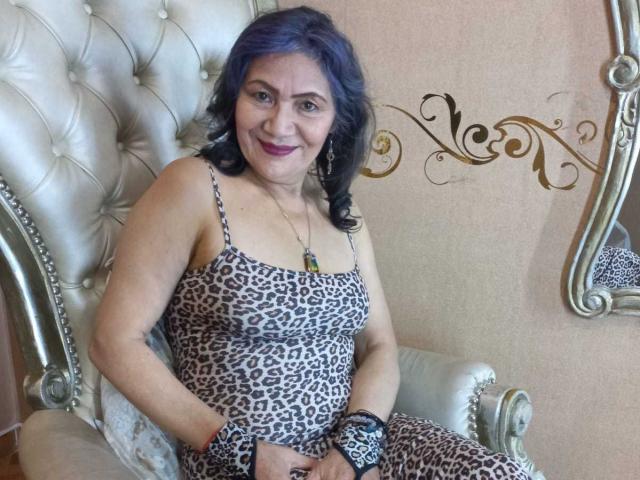 CelesteBakers with blue hair, leopard print dress, and purple lipstick, sitting on a silver tufted chair, smiling at the camera, wearing a black lace wristband.