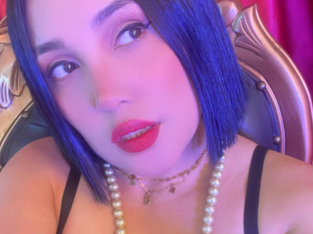 SamantaMuller with blue bob, red lipstick, pearl necklace, gold chain, black strap top, looking up, nose ring, pink-purple lighting, soft focus. Boobs visible, slightly out of frame.