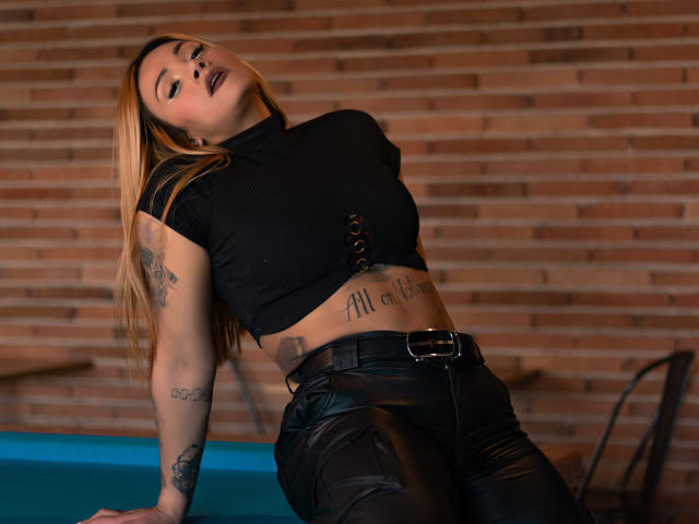 AzhunaStar with long blonde hair, black crop top, and black leather pants, leaning back, mouth open, tattoos on arms and stomach, brick wall background, blue table in front.