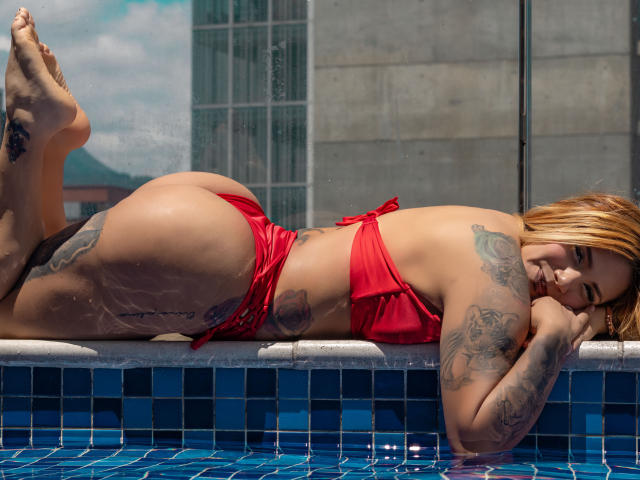 AzhunaStar lies by the pool, wearing a red bikini, her ass up, tattoos on her thighs and arms, blonde hair, smiling, water droplets on her skin, legs up.