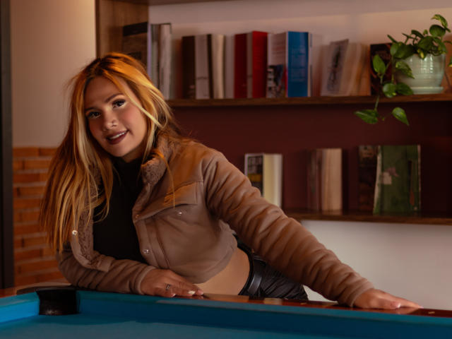 AzhunaStar with blonde hair, wearing a beige jacket, leans on a pool table, smiling, showing her ass, in a cozy room with bookshelves and a potted plant.