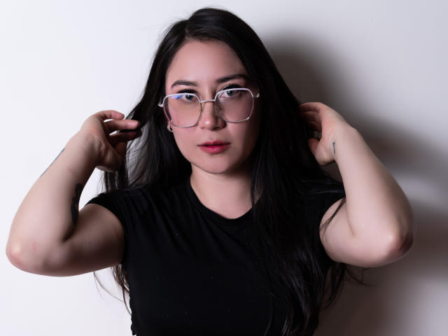 LeahMuniz with long black hair, glasses, and red lipstick, wearing a black shirt, hands in hair, fair skin, looking at camera, slightly smiling, soft lighting.
