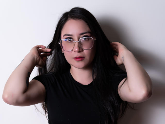 LeahMuniz with long black hair, wearing clear glasses, black shirt, hands in hair, fair skin, pink lips, looking at camera, neutral expression, slight tattoo on left arm.