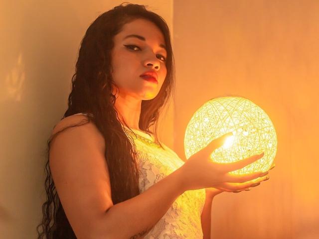 JenAndPatrick with long wet black hair, holding a glowing spherical light in her hand, wearing a white lace dress, standing against a warm orange wall.