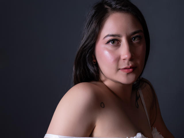 Leah Muniz tiene el cabello castaño oscuro, piel clara y un tatuaje pequeño en el hombro derecho. Lleva un sostén blanco, mostrando sus pechos, y tiene una sonrisa sutil..