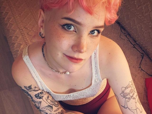 ShellyLove69 with pink hair, blue eyes, and tattoos, wearing a white lace tank top and red underwear, looking up with a slight smile. Her tits are small, and she's cute.