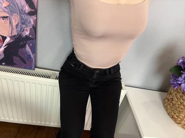 Amabel's pale pink tank top shows her big boobs, tight black jeans, and a slightly visible brown belt. Purple flowers in a woven basket on the right.