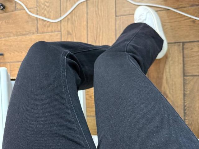Amabel's black jeans and white sneakers are in the frame, with her legs crossed and a white wire on the wooden floor.