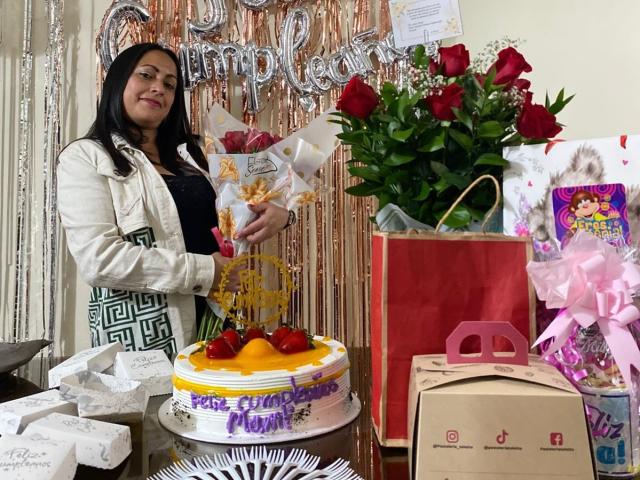 ScarlettBarness with black hair, wearing a white jacket, holds a bouquet of roses and a gift. She's standing by a cake with red cherries and a red bag.