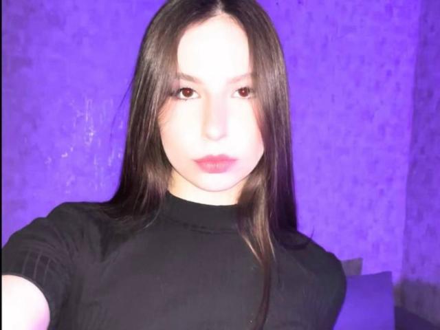 Amabel with long brown hair, wearing a black turtleneck, has pale skin and pink lips, looking directly at the camera in a purple-lit room. Her tits are not visible.
