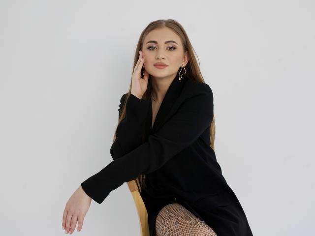 BeataAngel, blond, in einem schwarzen Mantel, sitzend, Hand am Kinn, Netzstrümpfe, Ohrringe, weißer Hintergrund, blickt in die Kamera, leichtes Lächeln, weiches Licht, stilvoll, sexy..