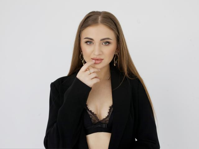 BeataAngel with long brown hair, wearing a black blazer over a black lace bra, has her finger on her lips, looking directly at the camera. Boobs visible.