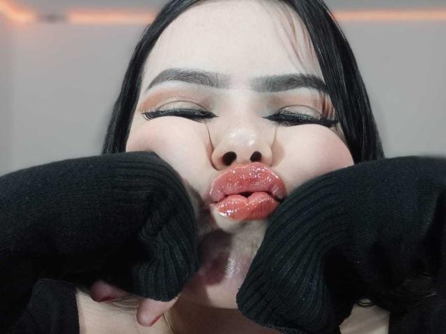 NathalyRose69 with glossy pink lips, black eyebrows, and black sleeves around her face, eyes closed, showing off her shiny lips. She has pale skin and straight black hair.