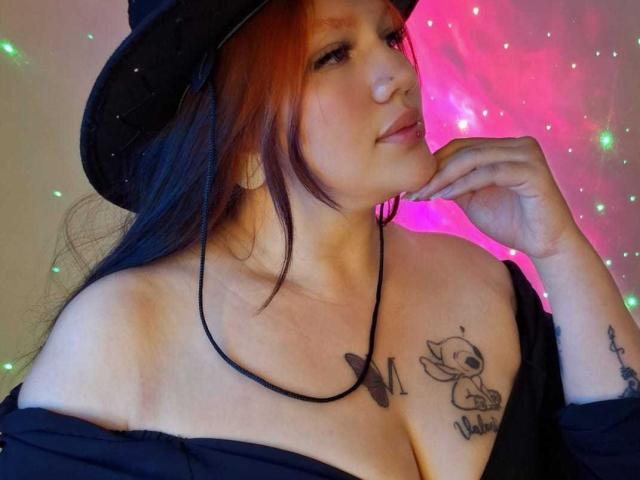 ValdemoreNm with long red hair, wearing a black hat, black off-shoulder top, showing a tattoo of Winnie the Pooh with "Maddie" on her tits, pink and green lights.