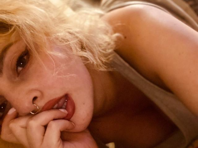 CocoVolkov with blonde curly hair, nose ring, and lip ring, lying down, tongue slightly out, hand near mouth, wearing a gray tank top, showing a bit of her tits.