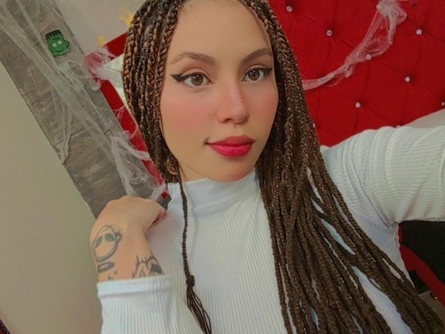 DominicTheOne has long brown braids, wearing a white ribbed turtleneck, with pink lipstick, and a tattoo on their right arm. Red headboard in the background.