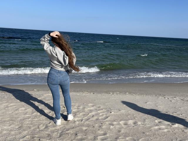 Dooris staat op een zandstrand, met haar rug naar de camera, en draagt een grijze hoodie, blauwe jeans en witte sneakers. Haar lange bruine haar wappert in de wind..