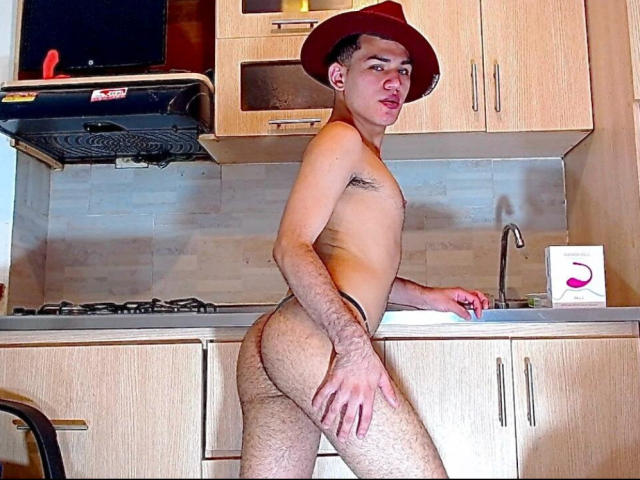 Alessandro Musk desnudo, con un sombrero rojo, de pie en una cocina, mirando de lado, mostrando su trasero peludo y sujetándose la mejilla. Su pene no es visible..