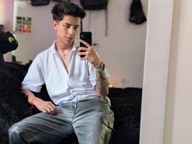 IanMonDumas, with short dark hair, white shirt, and gray pants, sits on black fur, holding phone, showing tattoo on right wrist, and looking at mirror selfie, showing a cute face.