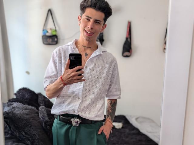 IanMonDumas with short dark hair, white polka-dot shirt, green pants, black belt, holding phone, smiling, tattoo on right arm, red bracelet, standing in bedroom, mirror selfie.