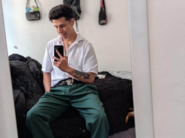 IanMonDumas, sitting, taking a mirror selfie, wearing a white shirt, green pants, belt, tattoo on left arm, smiling, holding phone, dark furry blanket behind, white wall, bags hanging.