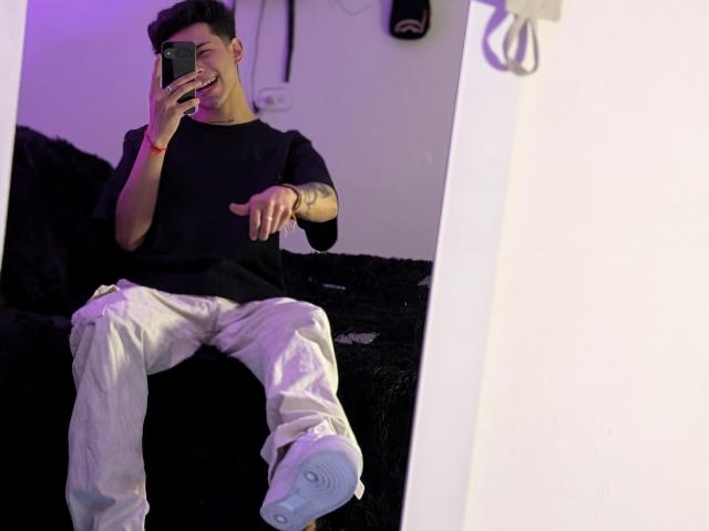 IanMonDumas, mirror selfie, black shirt, white pants, pointing, holding phone, black bed, white wall, white wall socket, purple light, short dark hair, tattoo on right forearm, white sneakers.