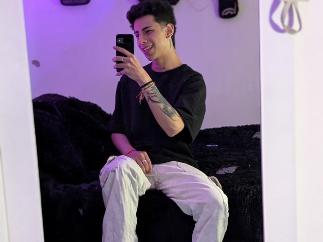 IanMonDumas with short dark hair, smiling, wearing a black shirt and white pants, holding a phone, tattoo on right forearm, sitting on a black fuzzy surface, mirror selfie.