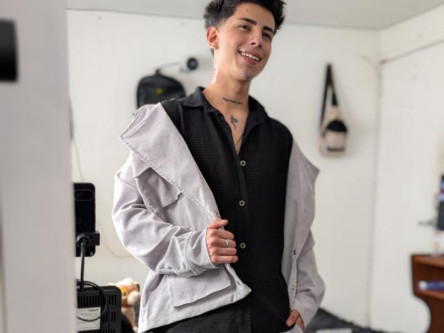 IanMonDumas with short black hair, smiling, wearing a black shirt and light gray jacket, holding the jacket's collar, has a star tattoo on his neck, standing in a room.