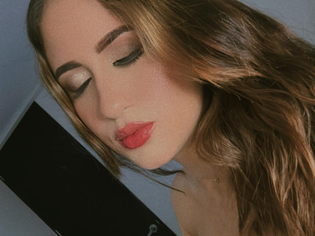 TiaraEvans has red lips, closed eyes with shimmering eyeshadow, and wavy brown hair. Her fair skin is slightly flushed, and she's wearing a delicate gold necklace.